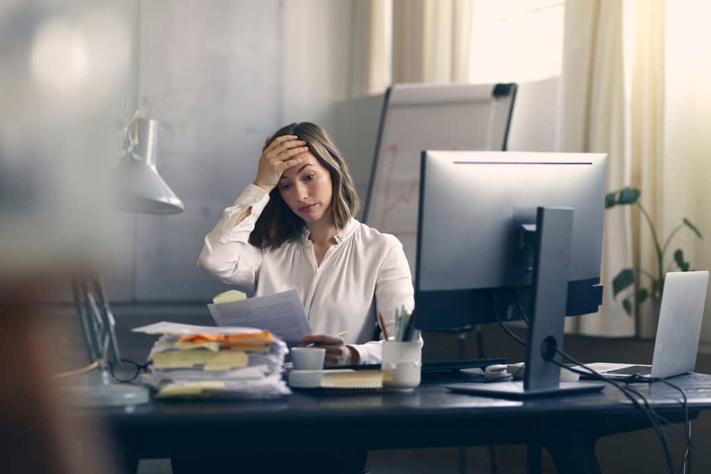 Very stressed business woman sitting in front of her computer looking at a large pile of paperwork
