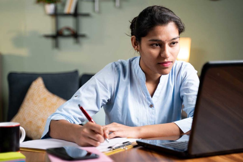 Young business woman writing down notes by looking laptop - concept of employee or student online training class at home.